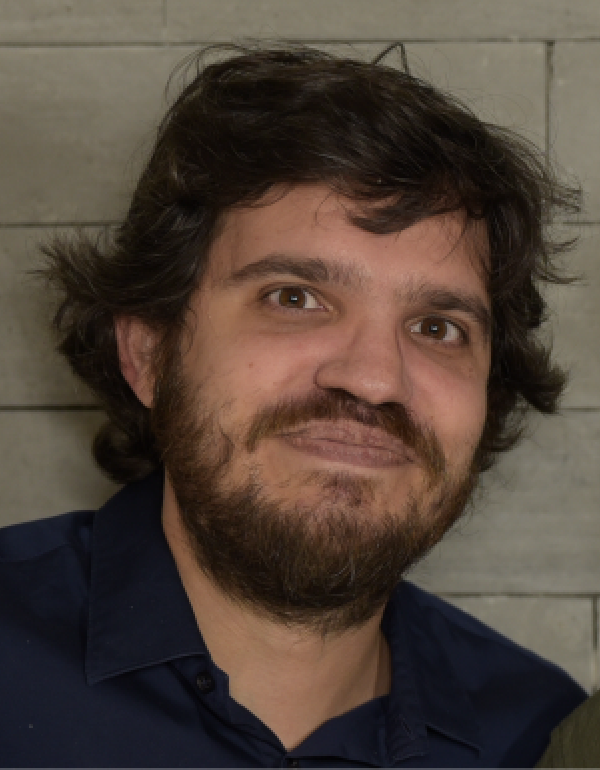A man with medium-length dark hair and a beard is facing the camera and smiling softly. He is wearing a dark blue shirt. The lighting is warm and even, showing his face clearly.