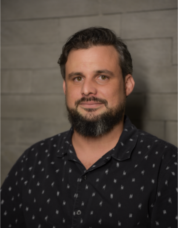 A man with medium-length dark hair and a beard is facing the camera and smiling softly. He is wearing a dark blue shirt.  The lighting is warm and even, showing his face clearly.