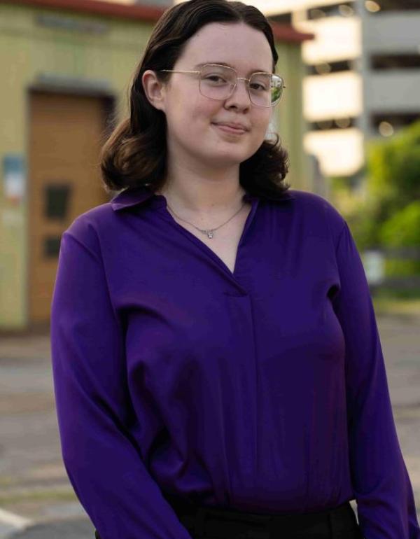 A pale 21 year old woman in a purple dress shirt and necklace. She has dark brown hair and square silver glasses.