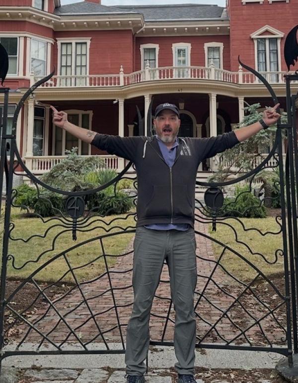 Man with hat standing in from of Steven Kings mansion gate that looks like spiderwebs. 