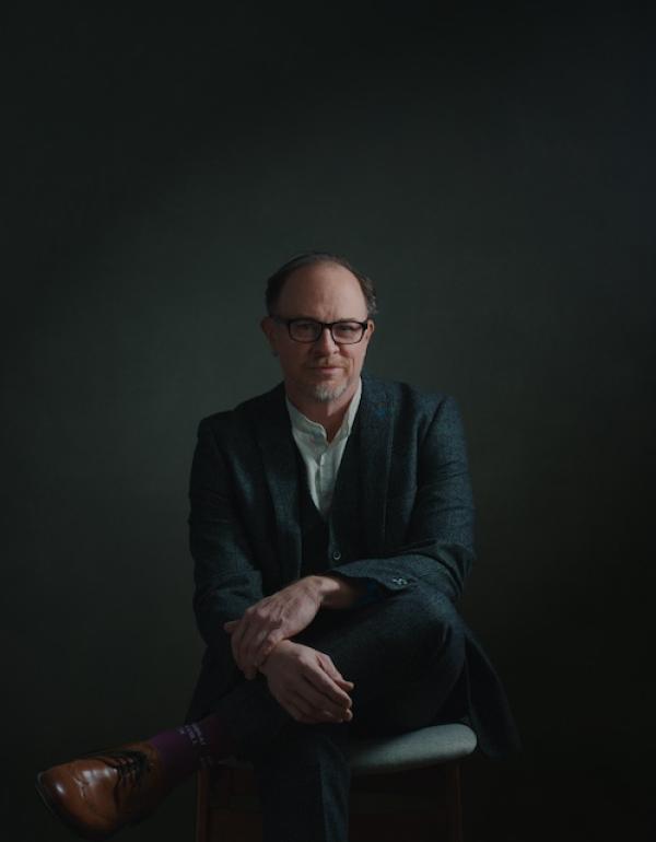 Chris Alan Evans sits cross-legged in a suit and glasses. Portrait by Alexander Jeffery.