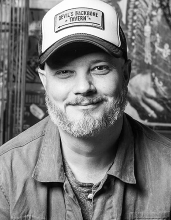 Black and white headshot of director Joshua David Matthews, wearing jacket and trucker hat saying Devil's Backbone Tavern
