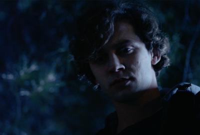 A young man stands under a tree, looking down at the ground with an inquisitive look in his eyes. 