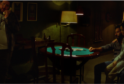 The scene shows a dark, cozy room lit by lamps. On the left, a heavyset man with a beard is standing and talking on the phone. In the center, there’s a green card table with playing cards and chips on it. On the right, a man sits holding a woman on his lap; both look serious and quiet. In the background, there’s a pool table and a green wall. The mood feels tense and silent.