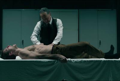 An older man in formal dress leans over a shirtless young man on a white-draped table in a cold, teal-lit room.