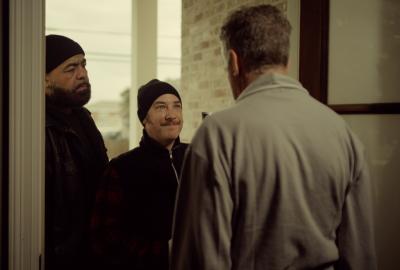 Two men in black knit caps stand at a front door speaking with an older man seen from behind inside a house.