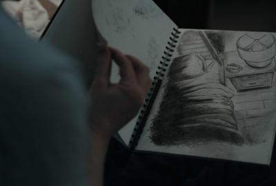 A woman's hand flips through a sketchbook showing a charcoal drawing of a bed and nightstand. A creature's creeps from beneath the bed.