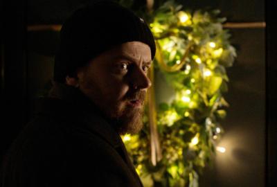 A bearded man in a hat looks back over his shoulder, an alarmed expression on his face. Behind him is a front door sporting an elaborate Christmas wreath.