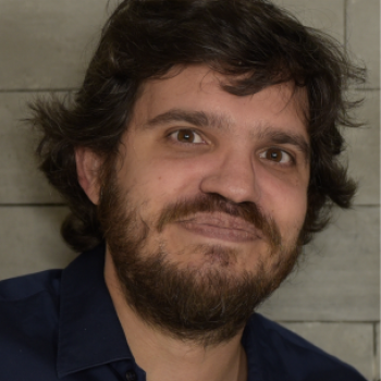 A man with medium-length dark hair and a beard is facing the camera and smiling softly. He is wearing a dark blue shirt. The lighting is warm and even, showing his face clearly.