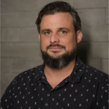 A man with medium-length dark hair and a beard is facing the camera and smiling softly. He is wearing a dark blue shirt.  The lighting is warm and even, showing his face clearly.