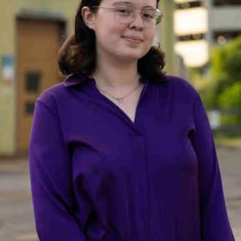 A pale 21 year old woman in a purple dress shirt and necklace. She has dark brown hair and square silver glasses.