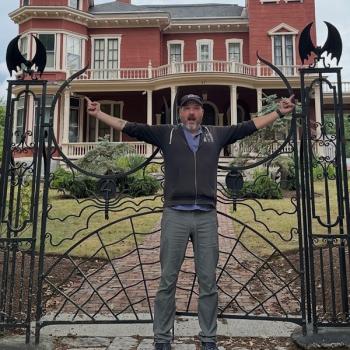 Man with hat standing in from of Steven Kings mansion gate that looks like spiderwebs. 