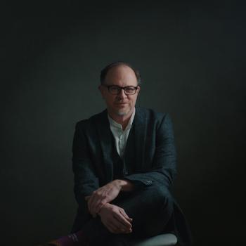 Chris Alan Evans sits cross-legged in a suit and glasses. Portrait by Alexander Jeffery.