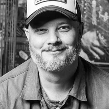 Black and white headshot of director Joshua David Matthews, wearing jacket and trucker hat saying Devil's Backbone Tavern
